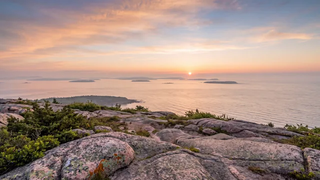 AI-created interpretation of sunrise from Cadillac Mountain in Acadia, with pink granite, island silhouettes, a calm Atlantic sea, and a peach sky.