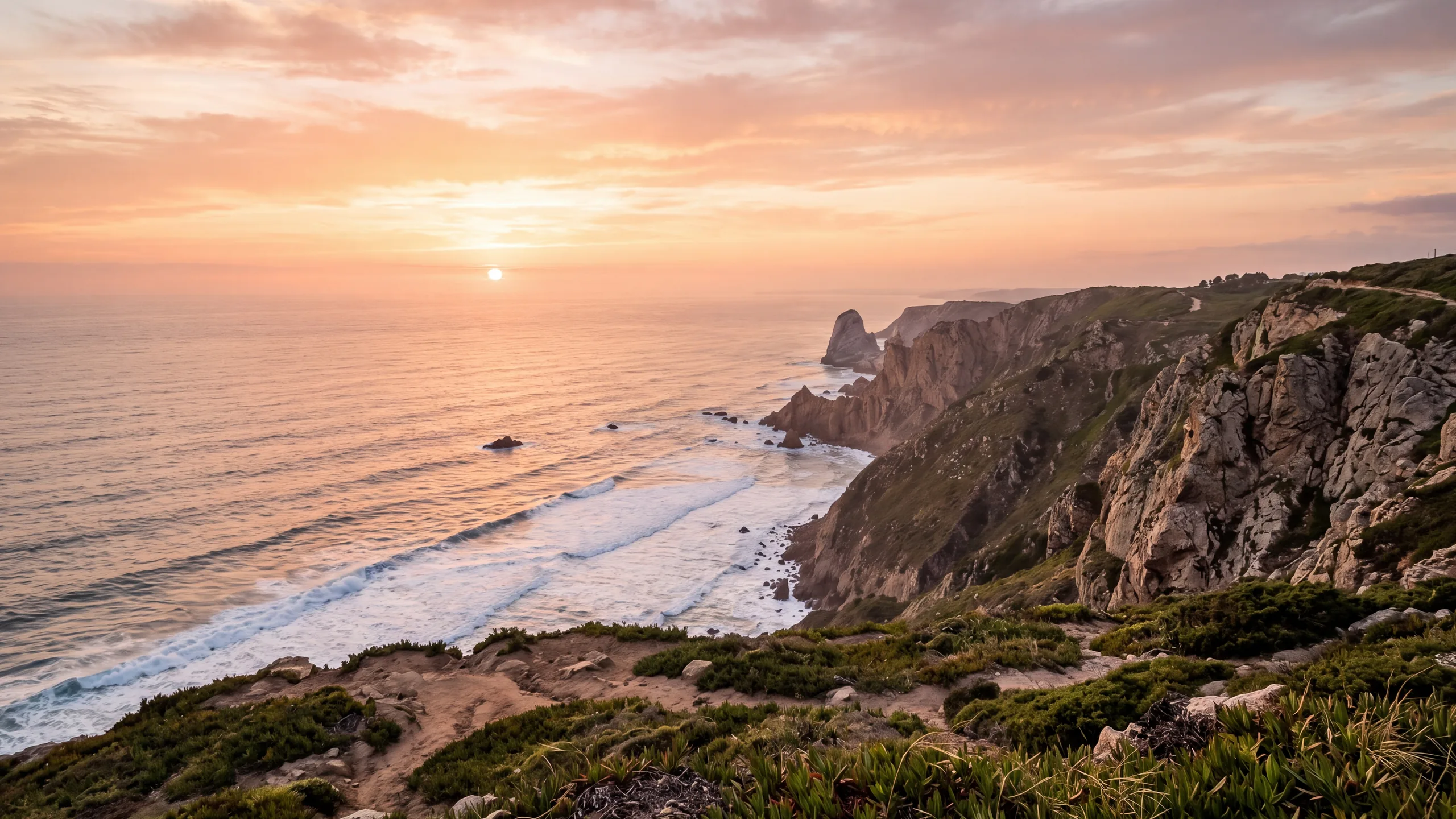 AI-created interpretation of Cabo da Roca on the Sintra coast of Portugal at sunset, with rugged cliffs, sea stacks, long white wave lines and coastal vegetation beneath a peach and amber sky.
