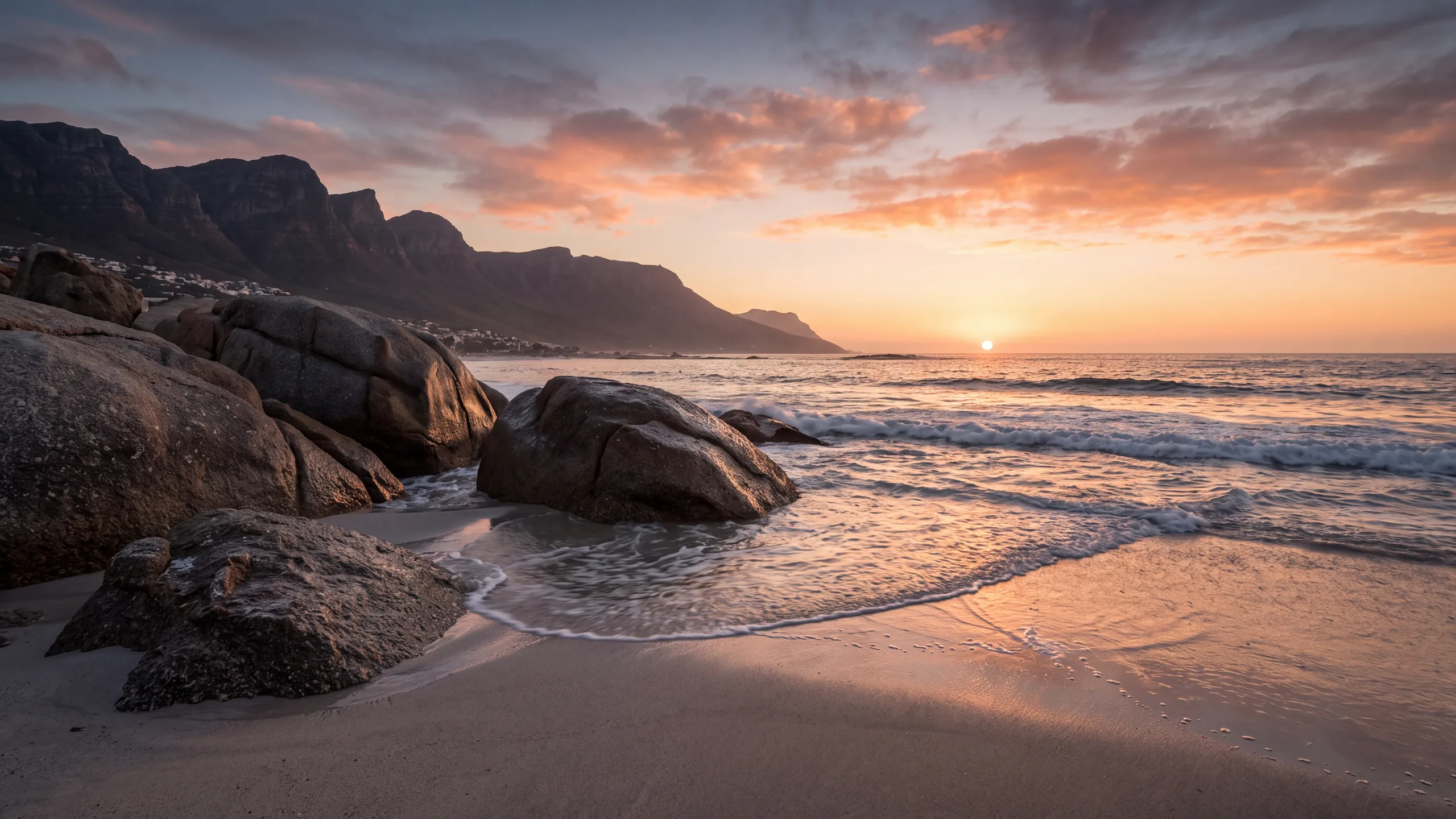 AI-created interpretation of Camps Bay in Cape Town, South Africa, at sunset, with granite boulders, retreating surf, a coastal mountain ridge and a small sun over the ocean beneath coral clouds.