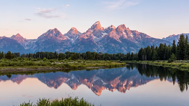AI-created interpretation of Grand Teton at sunrise, with rose-gold peaks reflected in a calm channel of the Snake River and a low fringe of green riverbank.