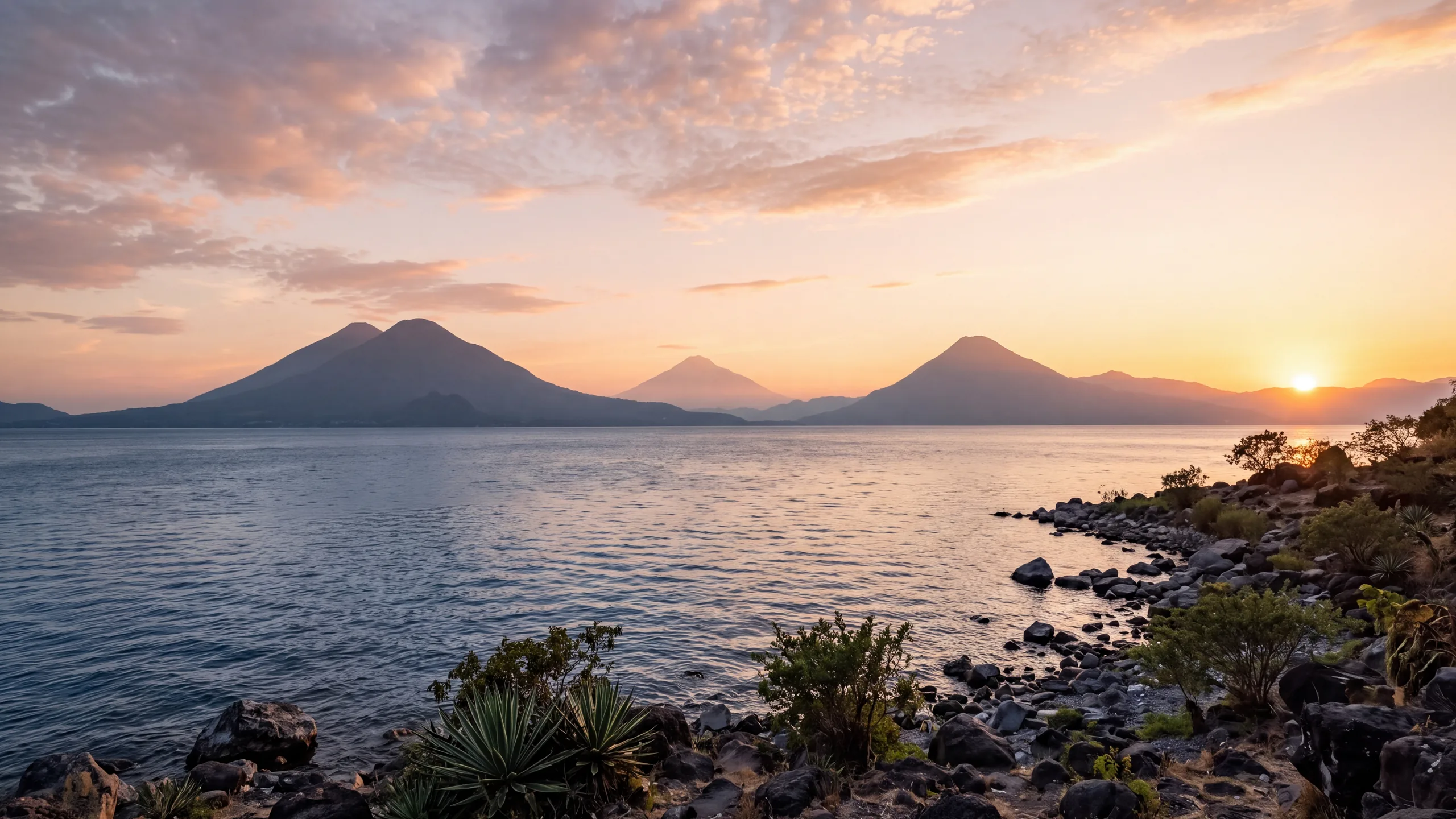AI-created interpretation of Lake Atitlán in Guatemala at sunset, with layered volcanic peaks across a blue lake, a rocky shoreline and coral clouds warmed by light from the right.