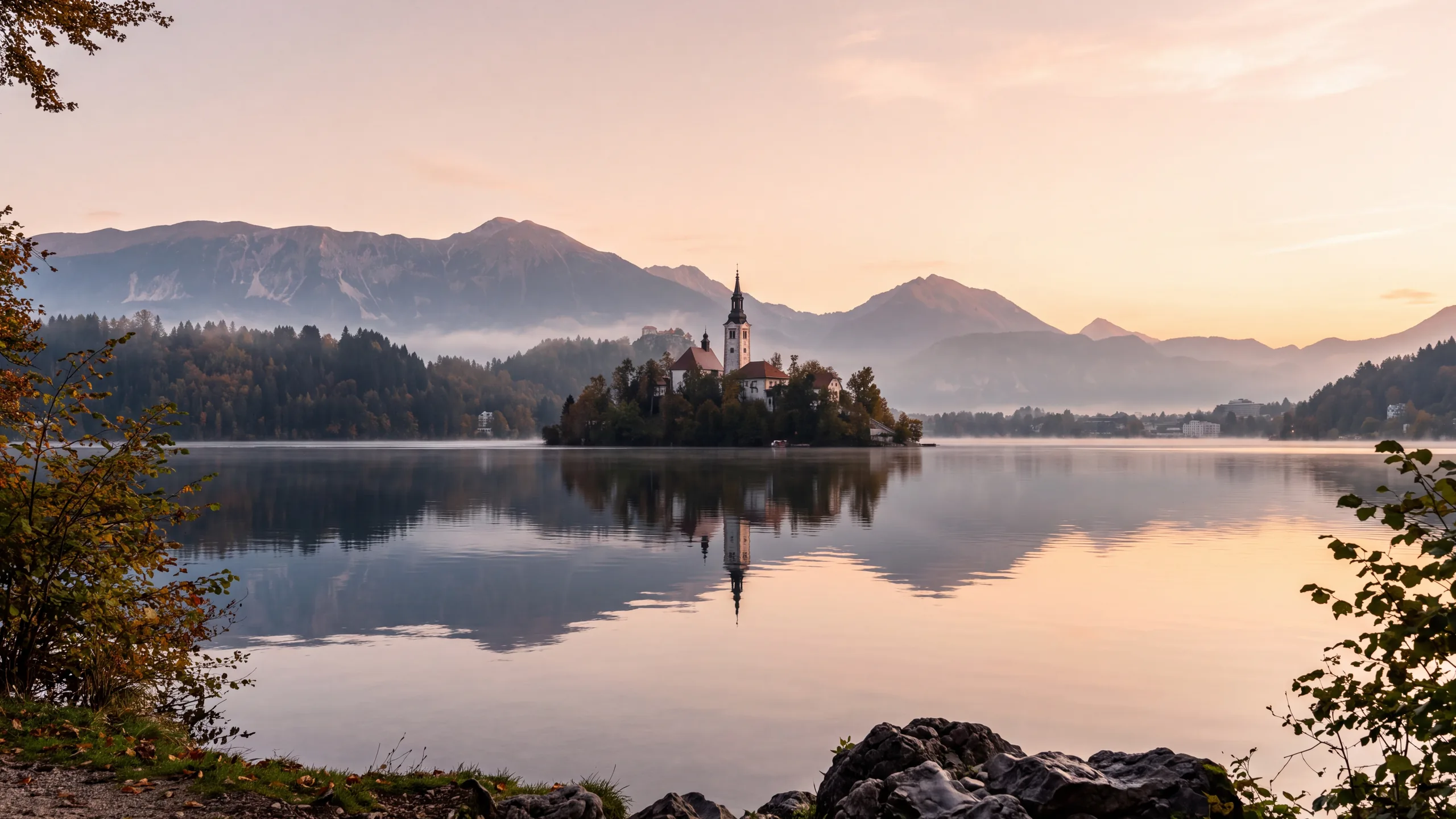 AI-created interpretation of Bled Island in Lake Bled, Slovenia, at sunrise, with a church reflected in calm water, misty mountains, soft peach light and autumn foliage.