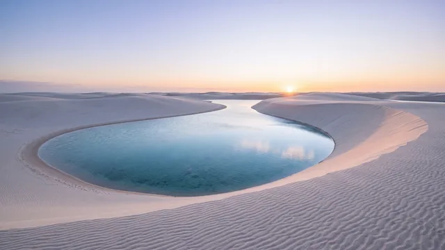 AI-created interpretation of Lençóis Maranhenses in Brazil at sunrise, with white coastal dunes enclosing a clear blue lagoon beneath a pale peach sky.