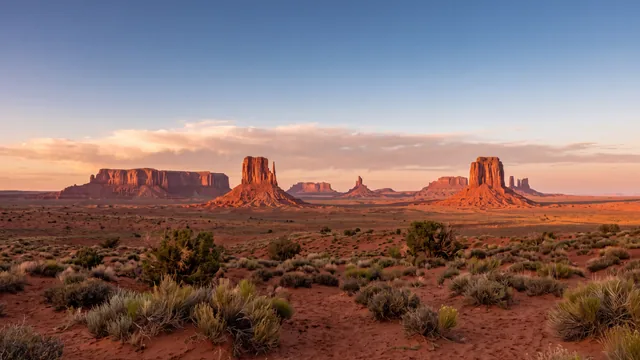 AI-created interpretation of Monument Valley in the Navajo Nation at sunset, with red sandstone buttes above a broad desert floor and soft coral clouds.