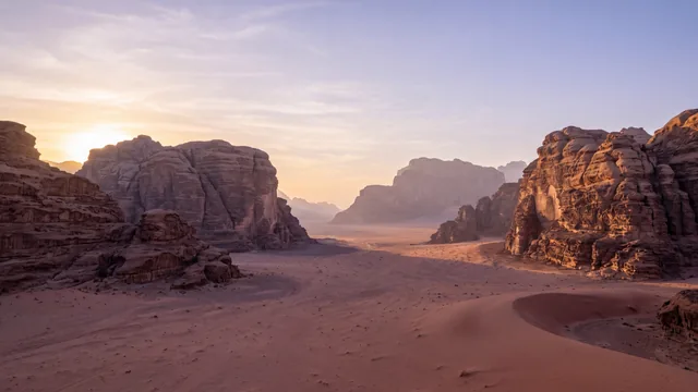 AI-created interpretation of Wadi Rum in Jordan at sunset, with red sand, weathered sandstone massifs and long soft shadows beneath a pale amber and lavender-blue sky.